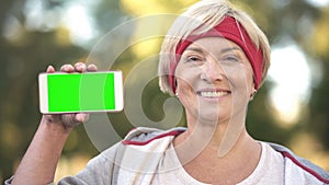 Smiling fit middle-aged woman showing green screen mobile phone, sports app