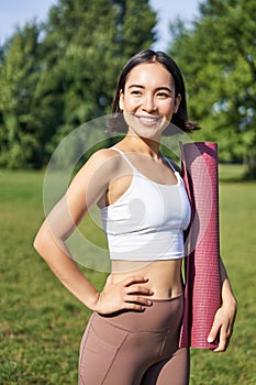 Smiling fitness girl with rubber mat, stands in park wearing uniform for workout and sport activities, does yoga