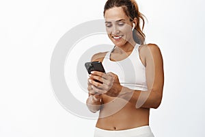 Smiling fitness woman looking at sport smartphone app, listening music wireless earphones, standing in activewear, white