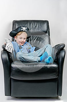 Smiling five year old boy dressed up in a pirate and police officer costume sit and laze in an black leather armchair.