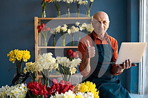 A smiling flower shop owner holds laptop in his hand and answers an SMS to client.