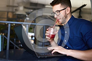 Smiling freelancer man with plastic cup of fresh beverage in hand works with laptop. Businessman in glasses drinks juice