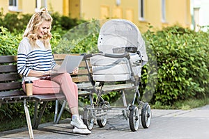 smiling freelancer working with laptop on bench near baby stroller