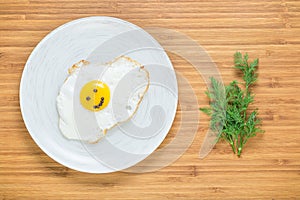 Smiling fried egg lying on a white plate on a wooden cutting board with bunch of dill. Classic Breakfast concept.