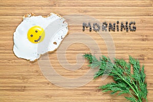 Smiling fried egg lying on a white plate on a wooden cutting board with bunch of dill and morning inscription near it.
