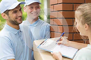Friendly couriers in blue uniforms and woman signing receipt of package delivery