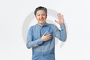 Smiling friendly-looking asian man being honest, raising arm and hold hand over heart while telling truth, making