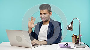 Smiling friendly man in business suit waving hand saying hisitting in office and talking to friend via web camera on his laptop