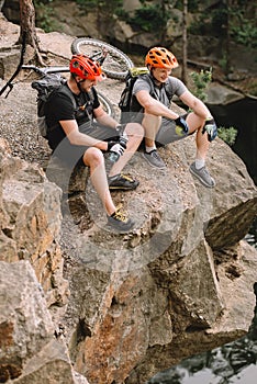 smiling friends cyclists with backpacks resting with sport bottle of water and apple on rocky cliff near river