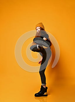 Smiling frolic red-haired girl in hat black tight jeans and boots is kicking over orange background