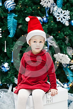 smiling funny Caucasian child girl toddler with blue eyes in red dress and Santa Claus hat sitting on gist box by New Year tree