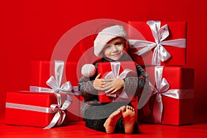 Smiling funny child boy in Santa red hat fell asleep with Christmas gifts in hand in hand on red background.