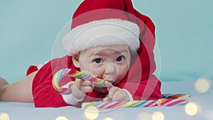 Smiling funny newborn baby in santa claus hat, red bodysuit play on white soft bed lick sweet. Concept holiday Christmas