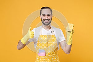 Smiling funny young man househusband in apron rubber gloves hold cleaning sponge rag while doing housework isolated on