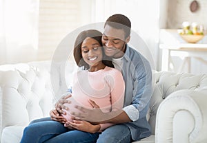 Smiling future mom and dad expecting baby, sitting on sofa at home and hugging