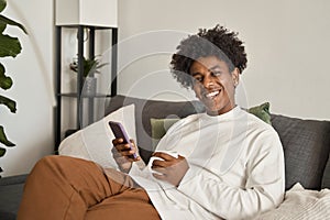 Smiling gen z African American teen sitting on couch using smartphone.