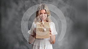 Smiling gentle romantic woman holding festive gift box opening desired surprise posing isolated gray
