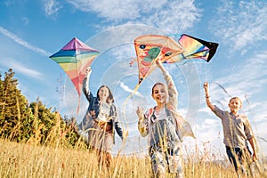 Smiling gils and brother boy running with flying colorful kites on the high grass meadow in the mountain fields. Happy childhood