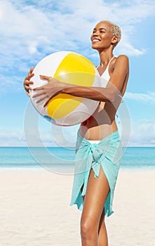 Smiling girl at the beach side holds inflatable beach ball, wearing a bikini and pareo, African latin American woman. Concept of a photo