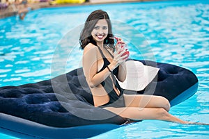 Smiling girl in black bikini holding a cocktail sitting on mattress in swimming pool on a blurred background of resort