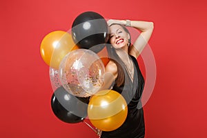 Smiling girl in black dress celebrate putting hand on head hold air balloons isolated on red background. St. Valentine`s