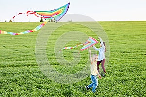 Smiling girl and brother boy running with flying colorful kites on the high grass meadow. Happy childhood moments