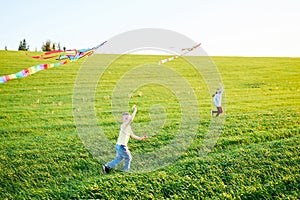 Smiling girl and brother boy running with flying colorful kites on the high grass meadow. Happy childhood moments