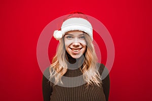Smiling girl in a Christmas hat and a warm sweater stands on a red background and smacks of the camera. Happy lady in santa claus photo