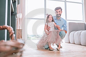 Smiling girl in cute dress sitting on father laps and looking at the camera in living room