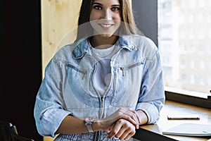 A smiling girl in denim clothes stands in front of the camera near the window and leans on the table.