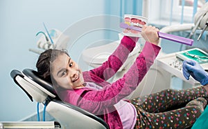 Smiling girl in dentist chair showing proper tooth-brushing using dental jaw model and big toothbrush in dental clinic