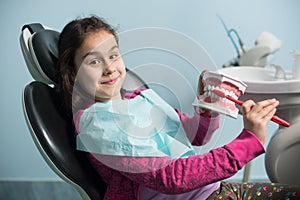 Smiling girl in dentist chair showing proper tooth-brushing using dental jaw model and toothbrush