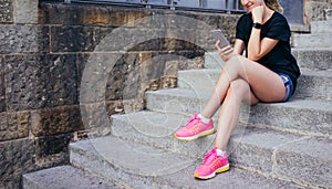 Smiling girl dressed in black T-shir, short denim shorts and pink sneakers sitting on stone steps and using smartphone