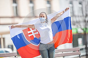 Smiling girl in a face mask holds a flag of Slovakia behind her on the street