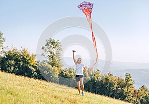 Smiling girl with a flying colorful kite running on a high green grass meadow in the mountain fields. Happy childhood moments or