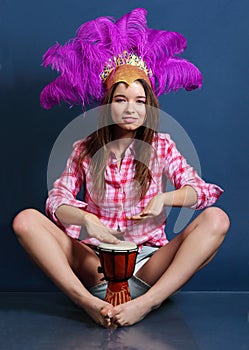 Smiling girl in hat with feathers sits on floor and beats drum