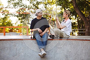 Smiling girl with headphones taking photo of young guy with skateboard on cellphone spending time together at modern