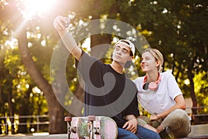 Smiling girl with headphones and young guy with skateboard happily taking photos on frontal camera of cellphone together