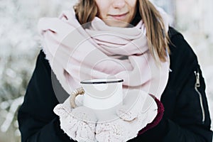A smiling girl holding a cup of tea on winter picnic in the woods. Woman hand`s in a warm pink knitted gloves
