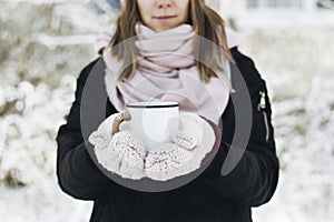 A smiling girl holding a mug of tea on winter picnic in the woods. Woman hand`s in a warm pink knitted gloves