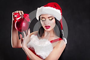 Smiling girl holding a red Christmas tree balls. Women on dress and santa`s hat. Santa`s helper .Attractive happy girl in santa h
