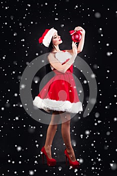 Smiling girl holding a red Christmas tree balls. Women on dress and santa`s hat. Santa`s helper .Attractive happy girl in santa h