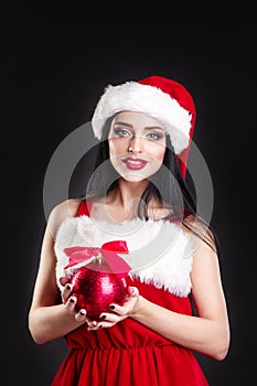 Smiling girl holding a red Christmas tree balls. Women on dress and santa`s hat. Santa`s helper .Attractive happy girl in santa h