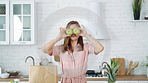 Smiling girl in the kitchen holds green apples in her hands and closes her eyes with them.