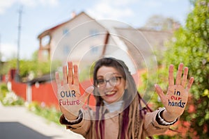 Smiling girl with long dreadlocks and eyeglasses in nude color trench is showing hands with written slogan `Our future