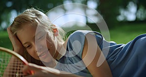 Smiling girl lying on green lawn alone. Blond kid touch racket net closeup