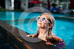 Smiling girl with pink goggles enjoys sunny day at outdoor swimming pool. Child relaxes in water, summer holiday fun