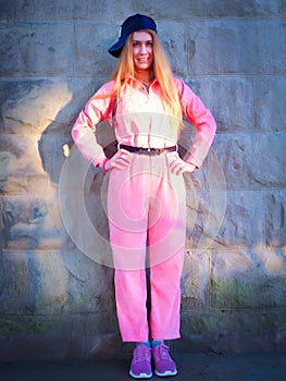 Smiling girl in pink jumpsuit stands by a stone wall in sunlight