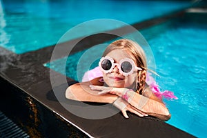 Smiling girl in pink swimwear, goggles enjoys pool time on sunny day. Kid relaxes on edge, cool water on summer vacation