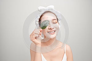 A smiling girl with a pink towel on her head applied a clay moisturizing mask while holding a leaf